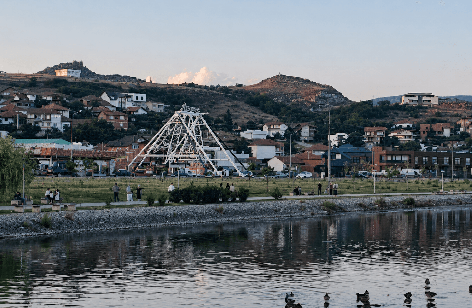 Daytime landscape across a river toward a hillside town, with a tall white lattice pyramid-shaped ride structure standing among the houses.